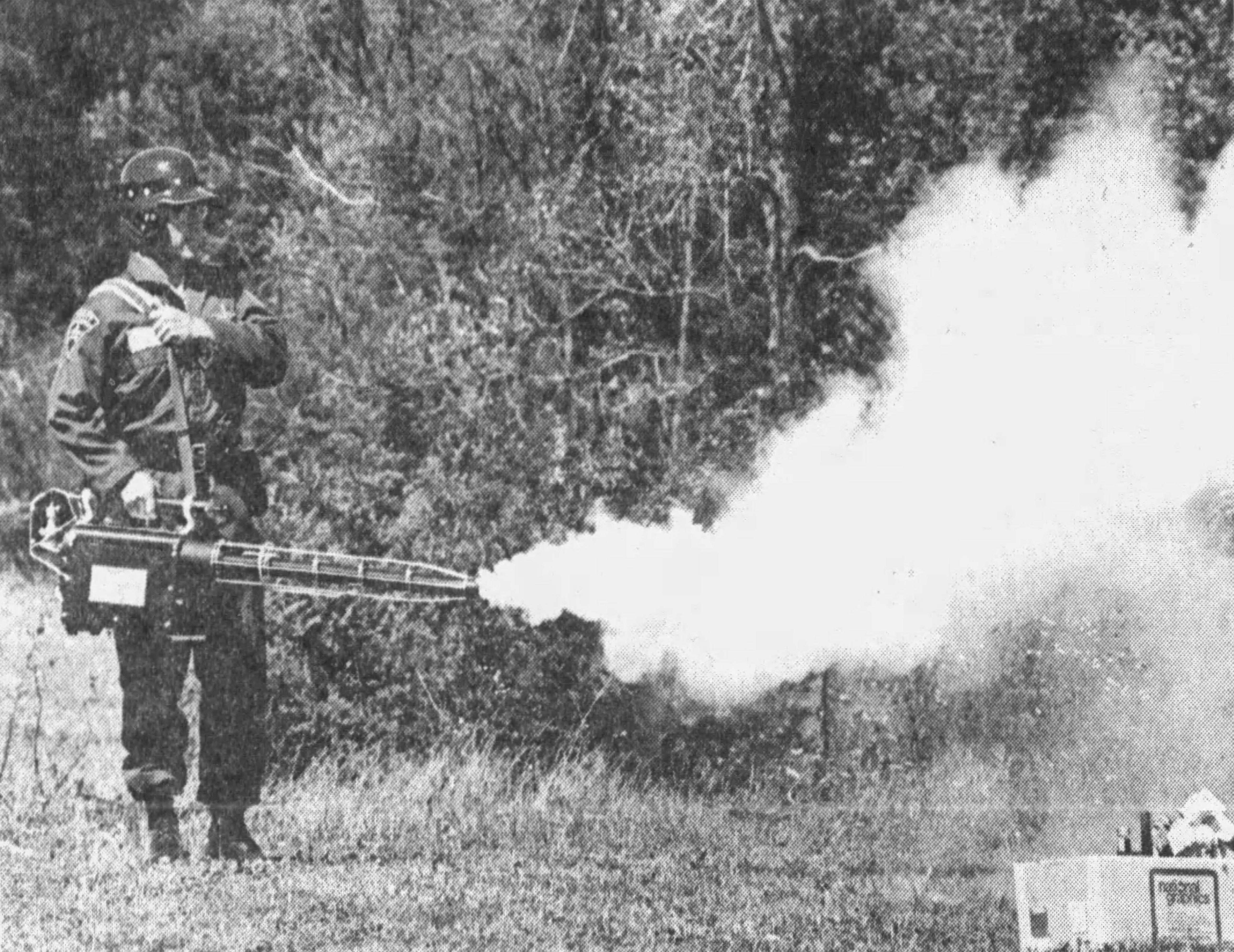 Black and white newspaper clipping of an officer standing in a field just front a forest/brush line, fogging out into the open area as part of a demonstration. The officer is wearing a riot helmet and coveralls and has the fogger slug over their right arm with a strap they are also holding with their left hand. The officer stands in the left part of the frame, fogging to the right, using a GOEC-style fogger with the nozzle tip right in the middle of the photo.