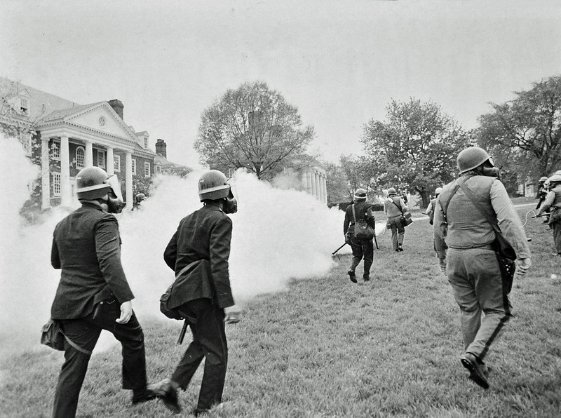 Black and white photo of a college quad with brick facade white column buildingsbehind trees around the left, back, and part of right sides of the frame. Eight or so police officers walking away from the camera wear riot gear. A few in dark, a few in light. One in light near the front is carrying a fogger with a shoulder strap and spraying a big cloud off to the left, obscuring a large portion of the photograph.