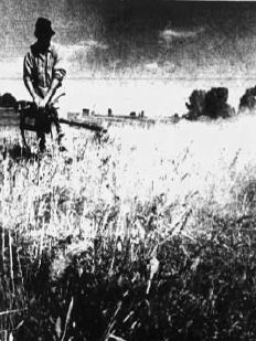 Black and white photo of an individual standing in a grass field with wood horse fence and trees and barns in the background. The individual is in light clothes and a black cap and is using both hands to hold a pepper fogger, which they are using to fog some grass on the right side of the photo. they are facing the camera, so the classic GOEC label is visible.