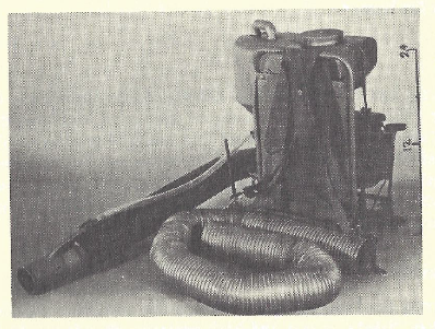 Yellowed black and white photo of a stationary Mighty Mite thermal fogger. It's a backpack fogger, so there's a giant hose that's like a vacuum hose wrapped around in the middle and then another one coming off of the actual backpack, which is upright in the middle back right. there is a metal frame and a large reservoir tank sitting on top of the engine and other aspects of the machinery. There is a tube running out to the end of the hose nose from the back pack. On the right side of the image is a scale bar that makes it seem like the backpack is 24 inches tall.