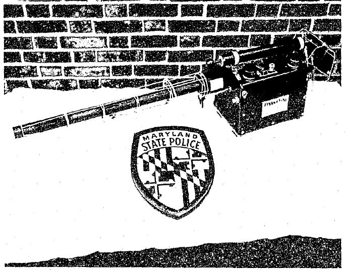 Black and white photo of a GOEC Pepper Fog fogger on a white ground with a maryland state police badge and in front of a brick wall. The fogger is in the mid right and points off to the left.