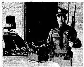 Black and white newspaper clipping of an officer standing in front of a open garage door, next to a police car that is partially in frame on the left and front areas. A GOEC-style thermal fogger sits on the hood of the car in front of the officer, pointing towards and to the left of the camera. The officer is wearing beat clothing and a cop hat and also has a shotgun.