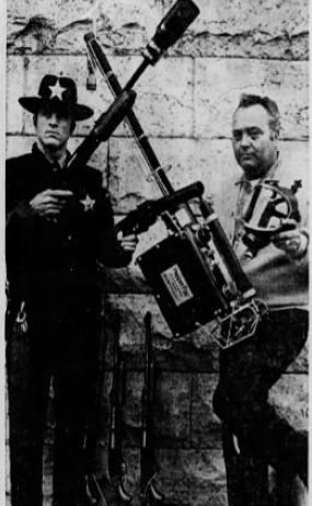 Black and white newspaper clipping of two officers standing in front of a large brick wall. Scott County deputy sheriff Jim Lewis, left, holds a new grenade launcher and a riot gun. he is donning a standard beat uniform with a bucket hat. Sheriff William Strout is on the right in street clothes and is holding a GOEC pepper fogger in his right hand and gas mask in his left. The officers are making an X with the barrells of the grenade launcher and fogger.