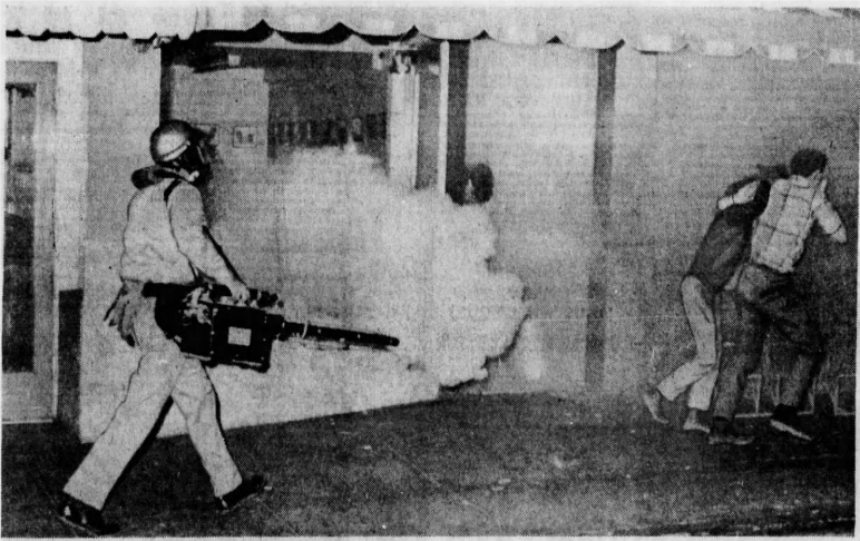 B/W newspaper clipping. To left is an officer wearing long pants, long sleeved shirt, and a helmet walking forward carrying a fogger in the right hand. The fogger is blowing fog through a tube and a cloud is forming. Background is a storefront window and door. To the right 2 people are moving away from the fog, leaning on one another, and covering their faces with their hands.