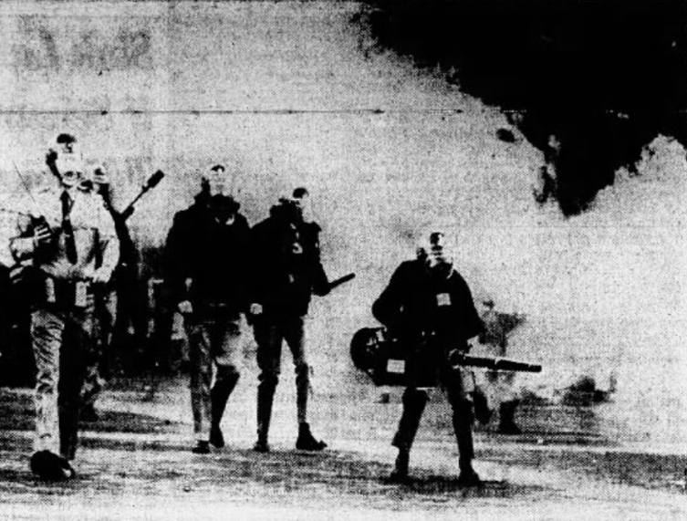 B/W newspaper clipping. Four people walking towards camera wearing helmets with face shields. Person on the left wearing white shirt and tie has several items hanging from belt. In right hand carrying radio with extended antenna. On right side person dressed in all black standing with a wide stance and holding pepper-fogger at hip height in right hand aimed forward. In center two more people dressed in all black, one with a short stick or club in left hand. Background is mostly cloudy with someone behind white shirt person, holding some sort of stick aloft.  Glimpses of additional bodies are visible in the cloudy background.    