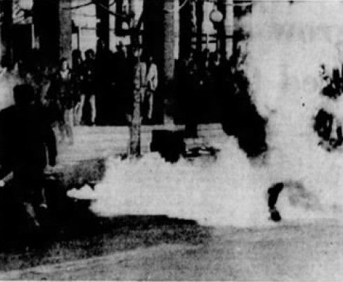 B/W newspaper clipping. Rather difficult to make out the people differentially in the background, but there's a large group of folks standing in the back part of the photo, the mid ground is relatively empty but filling with a cloud of white smoke eminating from an area on the left that appears to be an officer holding a thermal fogger. Behind everyone is the student union. 