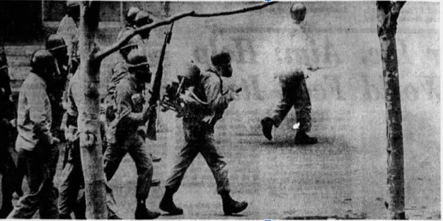 Guardsmen with bayonetted rifles and sheriff's deputies with tear and and pepper fogger walking through UC campus: B/W newspaper clipping:  Eight people in masks, shields, and tied boots walking from left to right, the one in front wearing a spray fogger strapped on back and holding the hose and nozzle in front. A cloud of fog is spraying from the nozzle. The second person is carrying a bayoneted rifle upright. The others are only partially visible as they are passing behind a tree. On the right ahead of the others an additional helmeted person can be seen turning towards the left. Two slim trees are in the foreground.