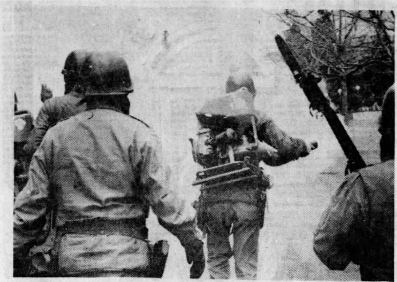 Four people walking away from camera, wearing helmets and holsters. Lead person has a fogger on their back and is holding the hose on the right spraying a fog ahead of them. Person on the right is carrying a bayoneted rifle raised above the left shoulder. The four are walking into the fog they’ve produced. There are some small trees to the right.