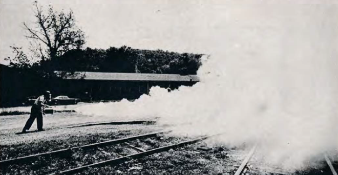 Black and white photo of a person using a pepper fogger across some railroad tracks. The person is standing in the mid ground on the left side of the photo and fogging towards the right mid-ground where the train tracks come from. The fog obscures the origins of the tracks off to the background on the right side of the photo. Behind the person on the left side is a taller tree along short building a car and some foliage. Further behind is a ridge of some kind with trees on it. The train tracks are old and partially overgrown.
