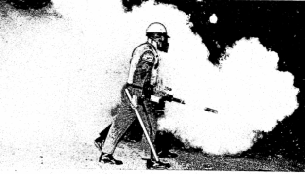 B/W Image: One person wearing gas mask and helmet, centered, stepping to the right. Person is holding slim white club in left hand and pepper-fogger in right hand. Fogger is pointing forward and a white cloud is surrounding the person to the front, back, and behind. The remaining background is black.