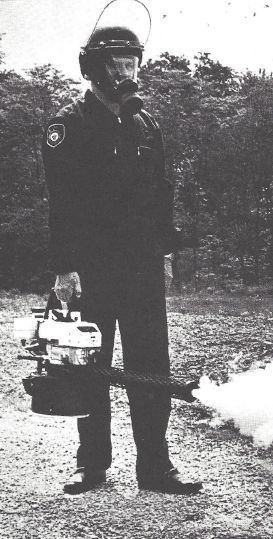 Black and white photo of police officer in a gas mask and riot helmet with the shield flipped up and full uniform, but not riot gear. The officer is holding a hand-held fogger that has a white top on the part, some shiny metal in the middle and then dark on the bottom with a dark nozzle that is spewing some fog. The officer is standing in a field in front of a forest.