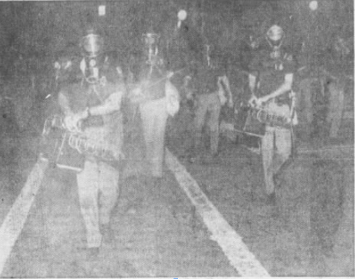 B/W image: Two people in foreground wearing helmets and face shields with gas masks and uniforms with short sleeves walking towards the camera, carrying boxy looking tools with nozzles pointing forward, with both hands. Person behind, also in short sleeve uniform, helmet, and gas mask carrying slim sabre or rod across the body. Behind these people seem to be more people but there are no clear details.