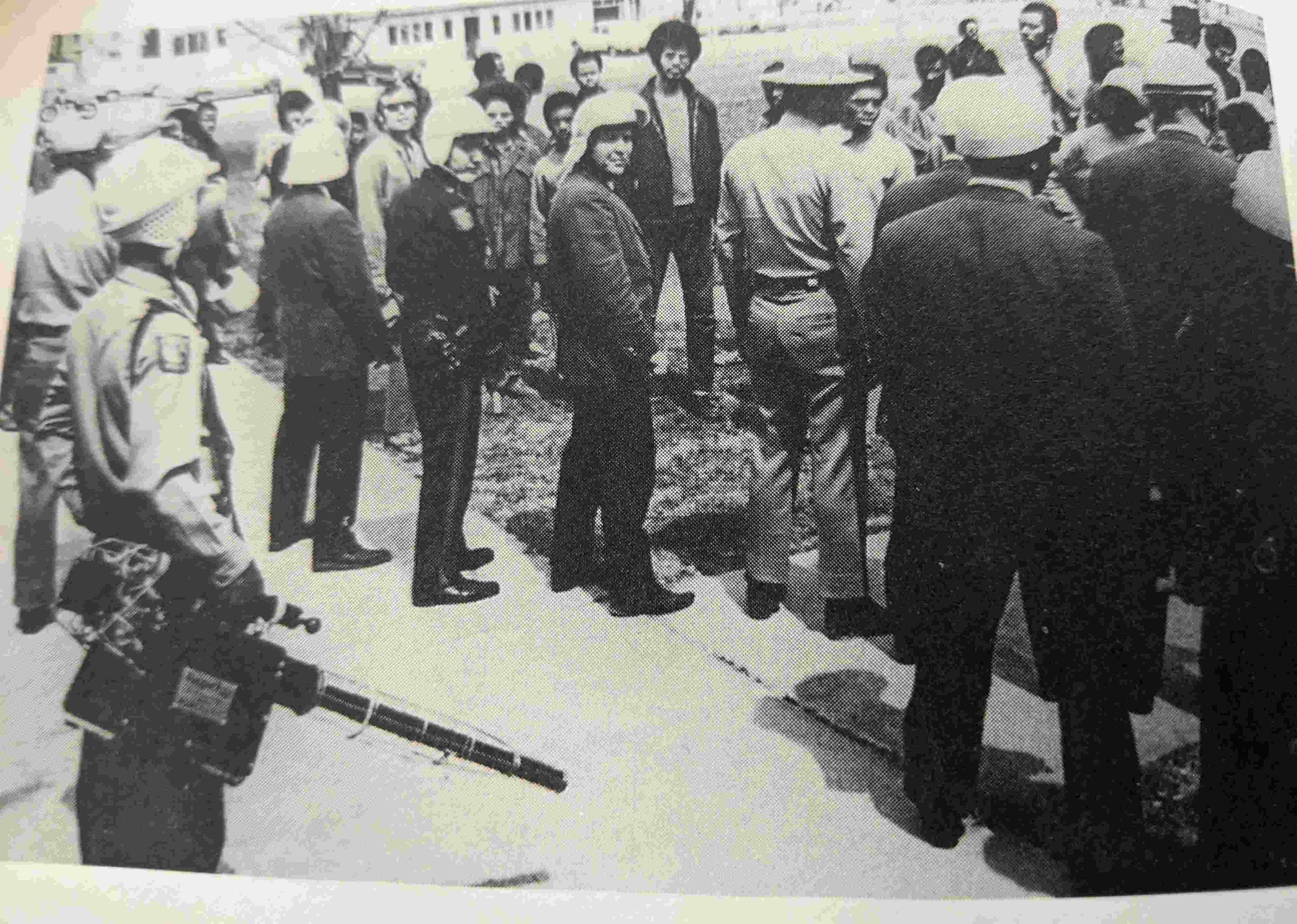 yellowed B/W faded image of police officers standing on a T of a sidewalk blocking the space from a group of predominately Black young people, who are standing behind them on the grass and facing the camera. Behind them are some cars and houses across a stree. The officer in the front left of the frame is carrying a Pepper Fog GOEC fogger.