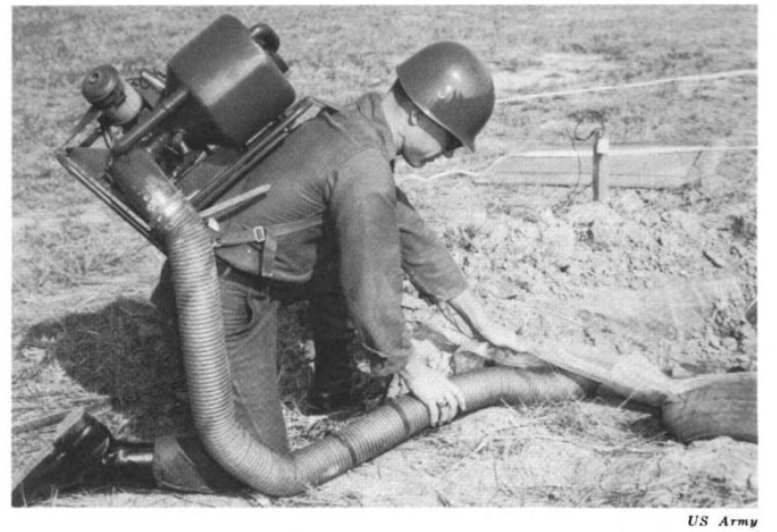 B/W image in a dirt field. Helmeted soldier on one knee with tank strapped on back. Lifting a board with left hand and holding an exhaust tube from the tank under the board with right hand.