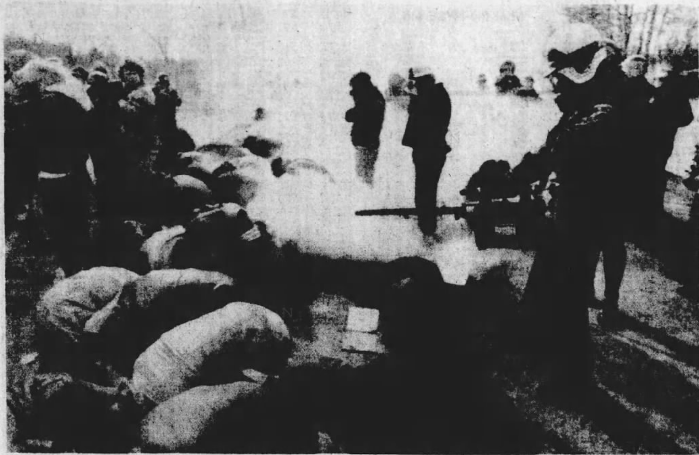 B/W newspaper clipping: To the left there are several people crouched on the ground with their heads down and covered. Behind them is a small crowd of people turning and moving away. To the right are three officials in helmets and masks facing the people on the ground and holding a fogger in front that is spraying a cloud of fog right over those on the ground.