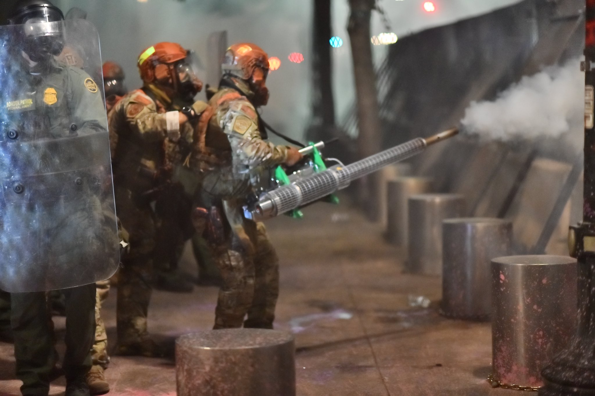 Fully riot-geared and for some reason in green camo US Homeland Security agents (to the middle and the left of the photo) behind a row of two-foot tall, one-foot radius metal posts, behind a metal grate wall over 7 feet tall with metal support beams and concrete pylon buttressing. In the front of the left side is an agent holding a plastic clear riot shield, through which you can see a patch that say 'Border Patrol Federal Agent' in yellow and some insignia patches as well. In the middle are the agents in camo, one with a hand on the shoulder of another who is operating a thermal fogger machine shooting gas through the fence. The machine is maybe four or five feet long and has a body not unlike a bush whacker with a two-cycle engine, but fueling a vaporizer instead of a rotor. The agent is holding the machine with their right hand visibly and there is a black strap across their shoulder holding it up. The machine is mostly shiny metal, although the tip is showing signs of corrosion (no surprise based on the compounds and heat) and the supports of the body are a bright green.
