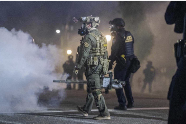 One person in green protective gear, wearing a bulletproof vest with weapons strapped to the body and wearing a helmet and gas mask is walking to the left carrying a fogger in the right hand arm extended down, nozzle pointing forward. A cloud of gas is coming from the nozzle. Next to them is someone dressed all in black with a bullet proof vest with the word POLICE across the back, also wearing a helmet and gas mask. It is night and there are additional clouds of gas and the shapes of people in the background.