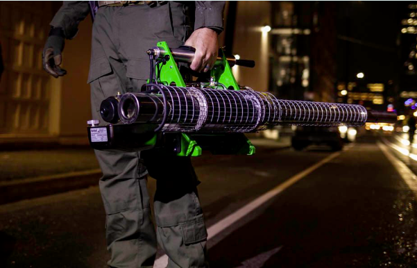 Night in a city, building lights in the background. One person standing alone in the center of a road, shown from the waist to the ankles. The person is wearing work pants with covered pockets at the thighs and calves, long sleeve shirt, and a glove on the right hand. In the left hand they are gripping the handle of a neon green fogger tool. The long black nozzle, covered with a wire cage, projects backwards and the motor is towards the front. It is being held at hip height; the arm holding it is relaxed down.