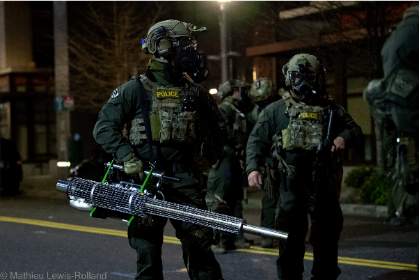 Night time with the light from a street light visible in the background. Two officers dressed in full protective gear with bulletproof vests holding supplies on, with the word POLICE stenciled in yellow. They are both wearing helmets and gas masks. The nearer one is holding a gas fogger in the right hand. Thefogger looks like a long tube between 3 and 4 feet long with a handle and motor parts near the back. The tube is covered with a wire cage until about the last half foot, which is a plain and narrower tube. Behind these two officers are some dimly lit buildings and one or two other officers but they are not clear.