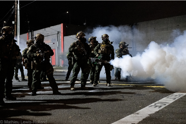 Nine people wearing full protective gear including helmets and gas masks standing spread out across a street at night. One is holding a gas fogger in one hand and gas is spewing and a cloud is forming in front of them. There is also some gas cloud behind the group. All of them seem to be wearing weapons on their gear but details are not clear. It is night. There is a grey building in the background with a red door and red trim. A white stripe on the roadway has the words MELT ICE spray painted on it.