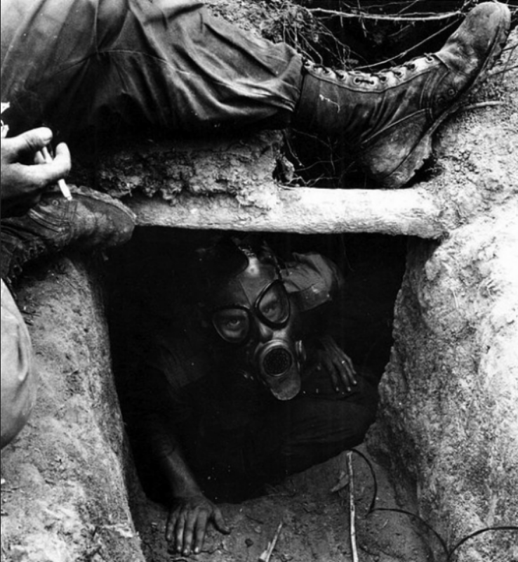 B/W image. Open trench at bottom, center. Pipe runs across trench and into the dirt on either side. Person in gas mask crouched below pipe looking up and forward. Leg in pants and lace up boot stretched over trench leaning against right edge. Other leg and boot partially visible on left. Hand holding lit cigarette resting on foot on left.