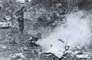 B/W image. In a clearing in a densely vegetated area, a small tank with an exhaust pipe blowing fog to the right. The cloud of fog covers much of the right side. Towards the back, 2 people wearing helmets and fatigues with sleeves rolled up stand with hands on hips on either side of the fogger, watching it.