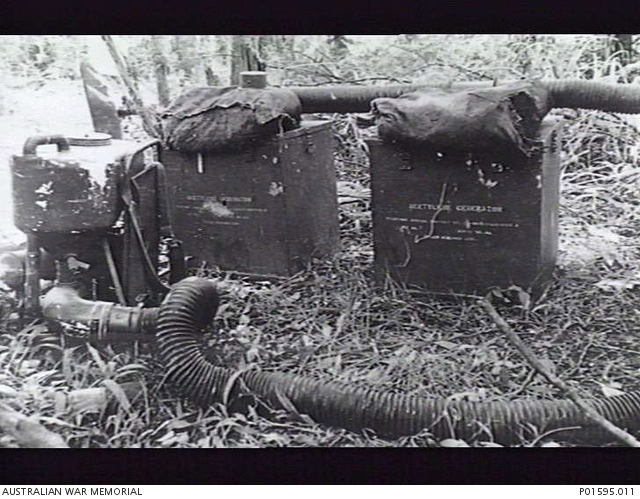 Black and white photo of a Mighty Mite blower on the left and two containers of acetylene in the middle, both containers are metal boxes with labeling in small white text and then some bladder bag on top. There are large vaccum size hozes coming off the blower and going off frame to the right. The scene is the ground of a jungle that has been cleared a little, there are trees and foliage in the background and dense but matted down grass in the fore.