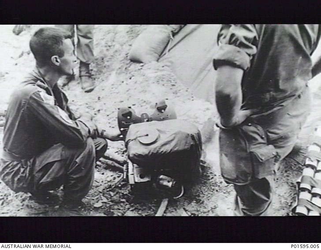 An individual crouches on the ground next to a blower, facing off to the right, with his left hand slightly resting on it. The photo is aimed down at this person, so the two people looking at the fogger while standing are partially visible from the feet upwards. The photo is an old black and white image and there are items around the sides that are difficult to make out, including potentially a cache of chemical weapons grenades on the right side and some sandbacks in the back.